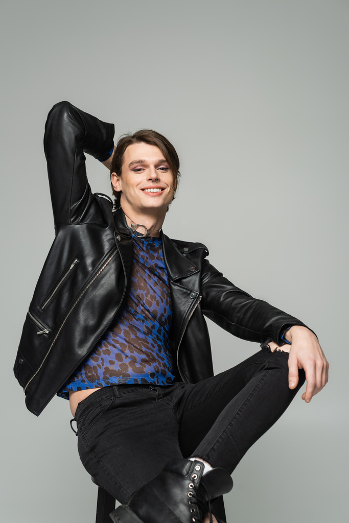 carefree bigender person in black leather jacket sitting with hand behind head isolated on grey - Photo, Image