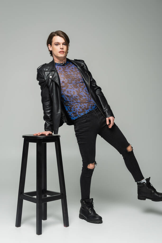 full length of bigender model in leather jacket and ripped pants posing near high stool on grey background - Photo, Image