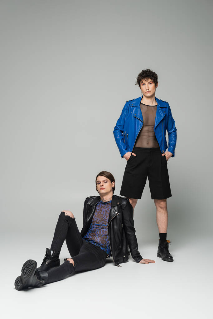 young nonbinary person in blue leather jacket standing with hands in pockets near trendy partner sitting on grey background - Photo, Image