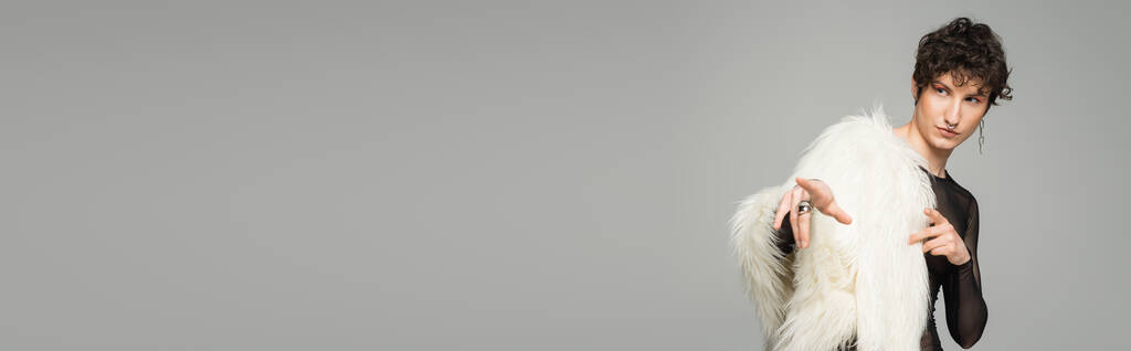 brunette bigender person in white faux fur jacket looking away and pointing with fingers isolated on grey, banner - Photo, Image