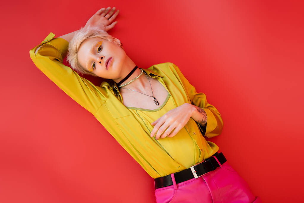 vista superior de la mujer albina de moda con delineador de ojos brillante y tatuaje mirando a la cámara en el fondo rosa carmín - Foto, imagen
