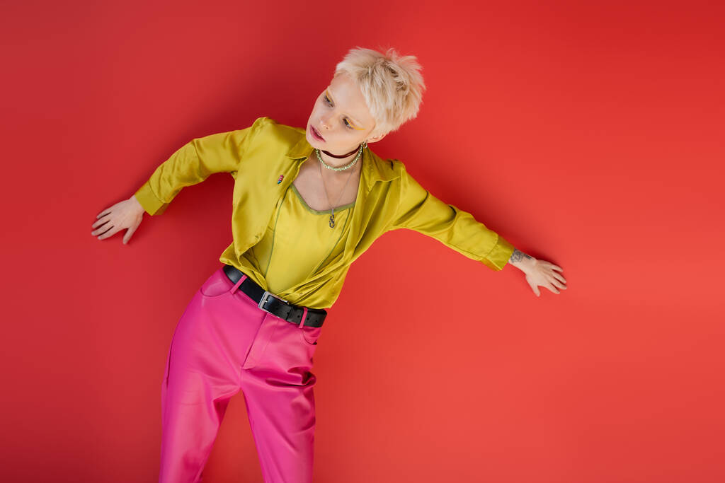 блондинка с светлыми волосами и татуированной рукой позирует в модной одежде на кармине розового цвета  - Фото, изображение