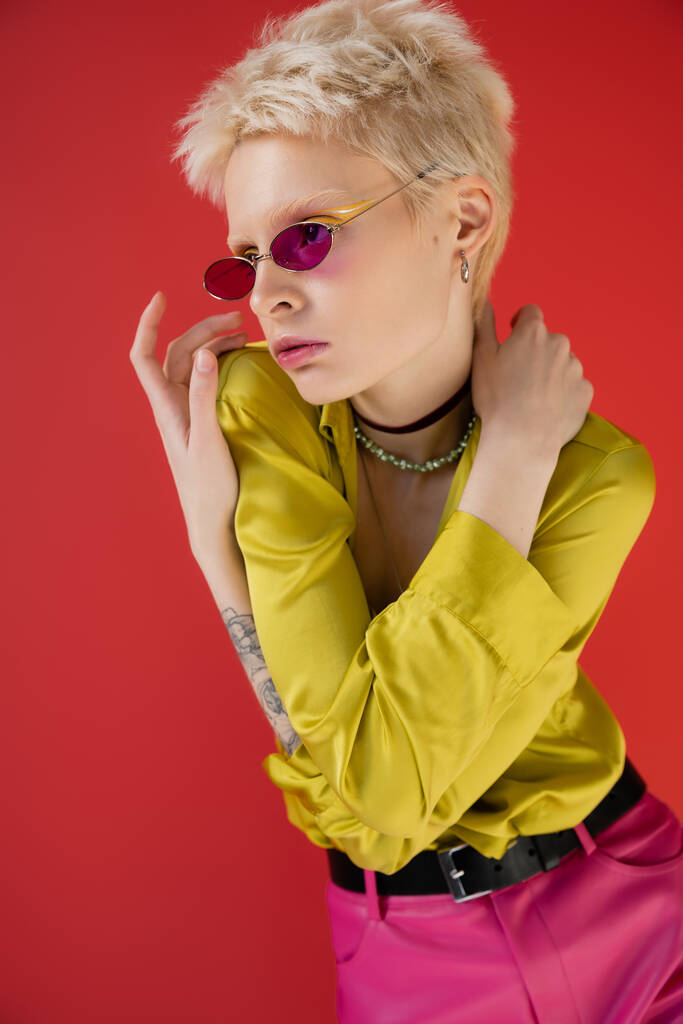 albino model with tattooed hand posing in stylish outfit and trendy sunglasses on carmine pink  - Photo, Image