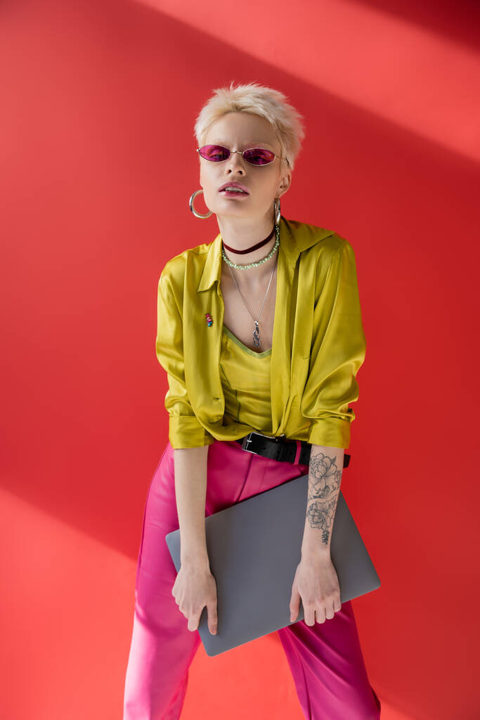 rubia albino freelancer con tatuaje posando en gafas de sol de moda y sosteniendo portátil en carmine rosa  - Foto, imagen