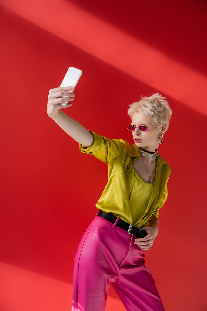 tattooed albino woman in stylish sunglasses taking selfie on smartphone on carmine pink background  - Photo, Image