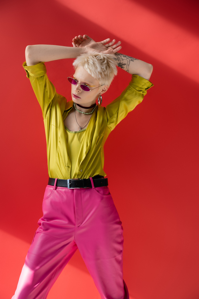 ξανθό μοντέλο με τατουάζ στο χέρι ποζάροντας σε μοντέρνα μπλούζα και μοντέρνα γυαλιά ηλίου σε καρμίνα ροζ  - Φωτογραφία, εικόνα
