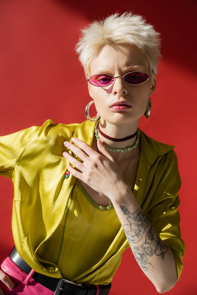 luz solar no rosto da mulher albina com tatuagem na mão posando em óculos de sol elegantes em rosa carmim  - Foto, Imagem