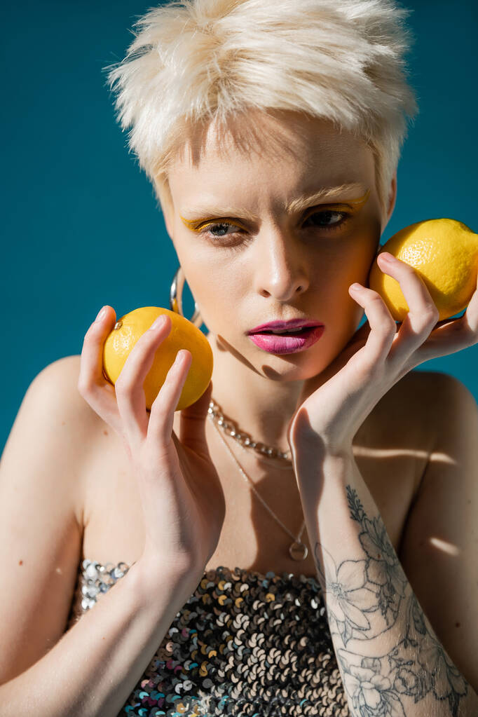 modèle albinos tatoué avec maquillage à la mode et cheveux blonds posant avec des citrons sur bleu  - Photo, image