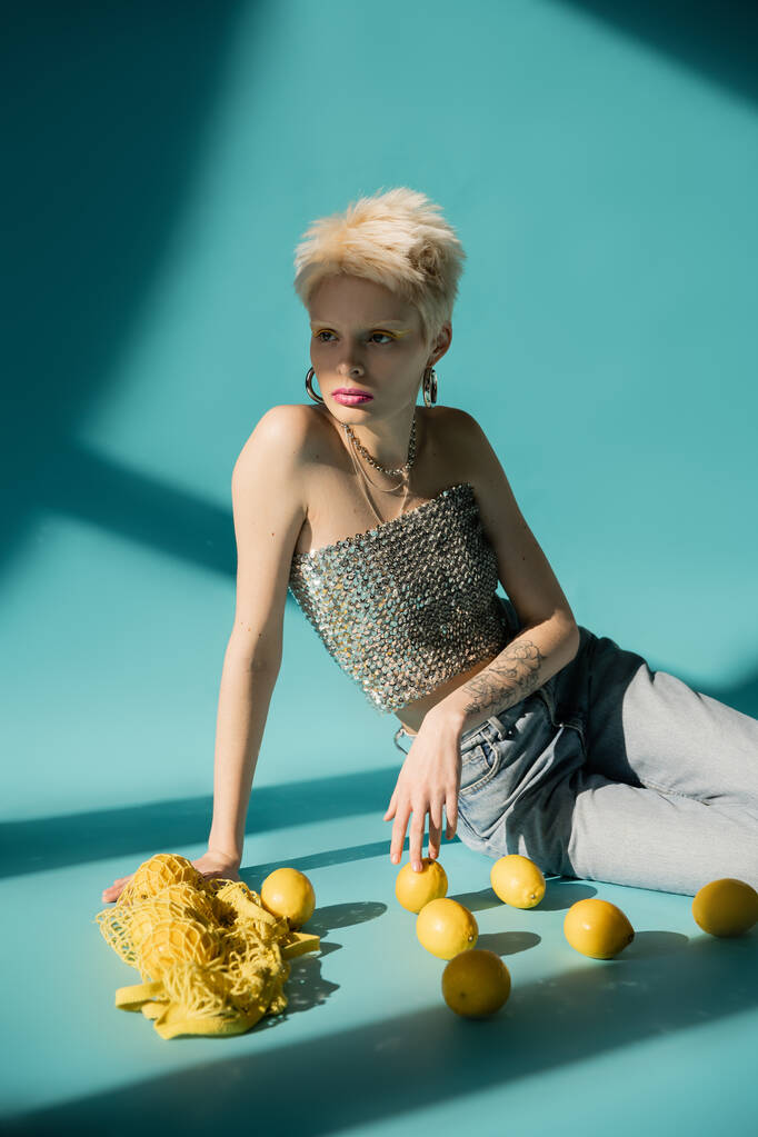 татуированная модель альбиноса в блестящем топе с блестками и джинсами, сидящими рядом с спелыми лимонами на голубом  - Фото, изображение