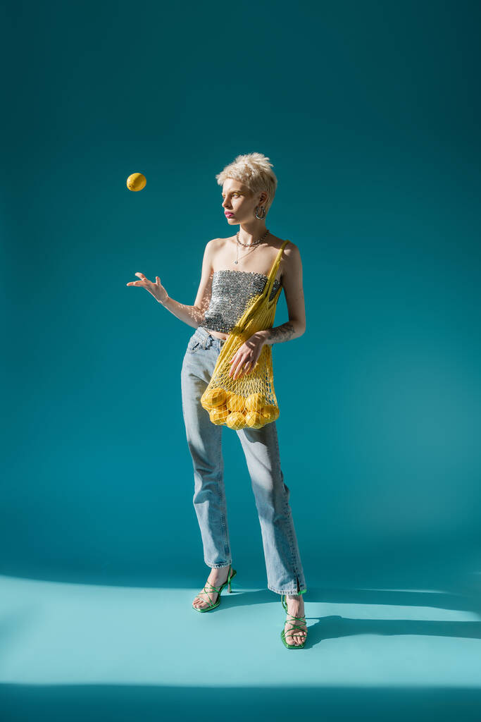 full length view of tattooed woman in shiny top with sequins holding net bag and throwing in air ripe lemon on blue  - Photo, Image
