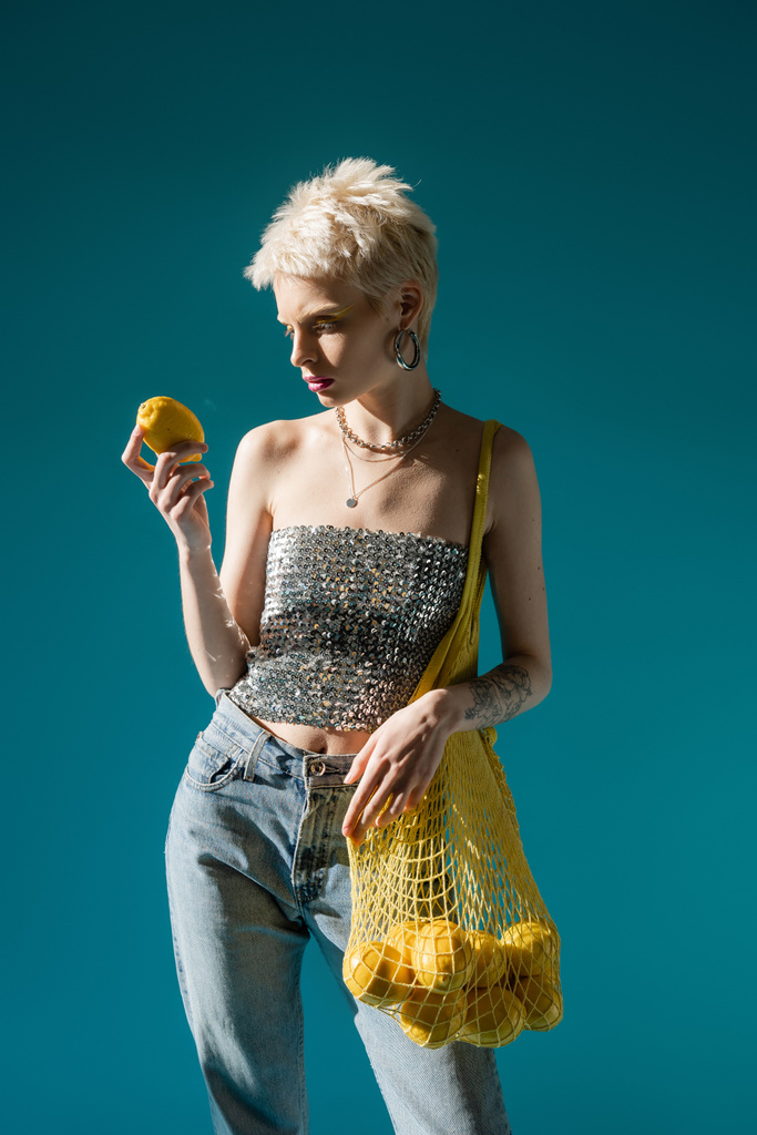 tätowierte Albino-Frau in glänzendem Top mit Pailletten, Netztasche mit reifen Zitronen auf blauem Grund  - Foto, Bild