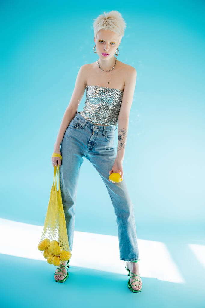 full length view of tattooed woman in shiny top with sequins holding yellow string bag with ripe lemons on blue  - Photo, Image