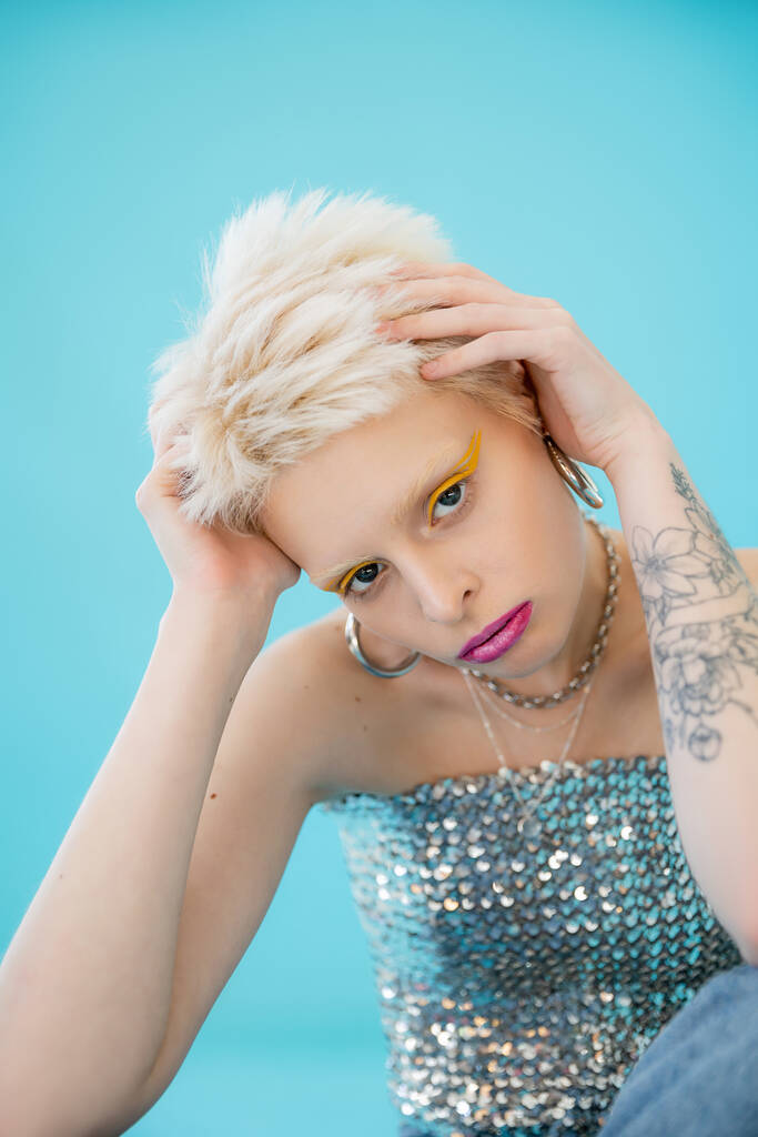 modelo albino tatuado em top brilhante com lantejoulas olhando para a câmera em azul  - Foto, Imagem