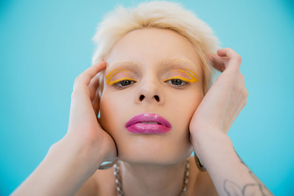 modelo albino loira com maquiagem brilhante olhando para a câmera no fundo azul  - Foto, Imagem