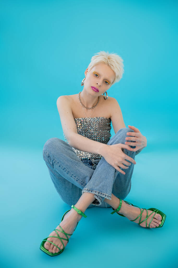 pleine longueur de blonde albinos femme avec des sandales à talons et jeans assis sur bleu  - Photo, image
