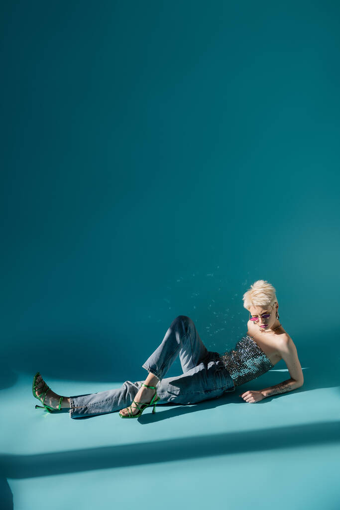 Pembe güneş gözlüklü, topuklu sandaletle mavi üzerine poz veren tam boy albino modeli.  - Fotoğraf, Görsel
