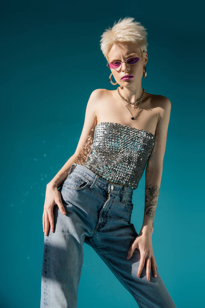 татуированная модель альбиноса в модных розовых солнцезащитных очках и модном наряде, позирующем на голубом  - Фото, изображение