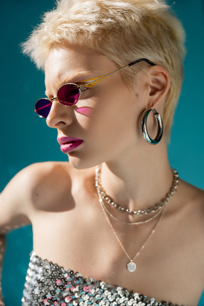 Porträt des blonden Models mit stylischer pinkfarbener Sonnenbrille und Schmuck posiert auf blauem Grund  - Foto, Bild