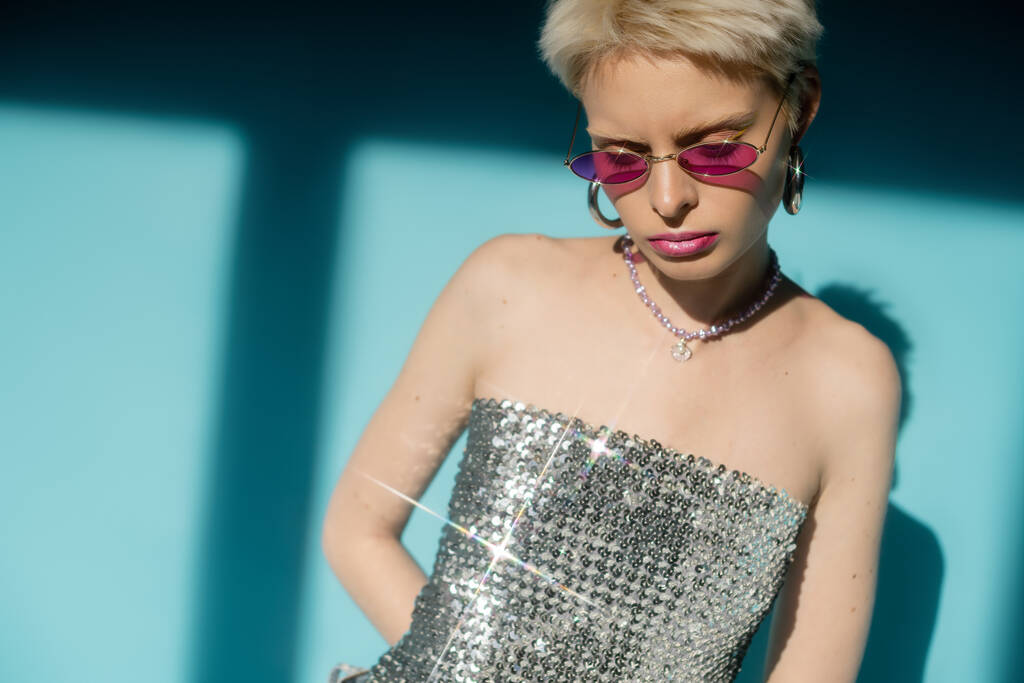 sunlight on body of albino model in pink sunglasses and shiny top with sequins on blue background  - Photo, Image