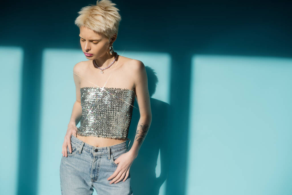 sunlight on albino woman in shiny top with sequins and jeans posing on blue background  - Photo, Image