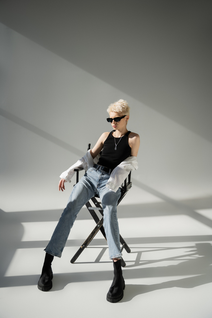 volledige lengte van stijlvolle blonde vrouw in zonnebril en trendy outfit zittend op klapstoel op grijs  - Foto, afbeelding