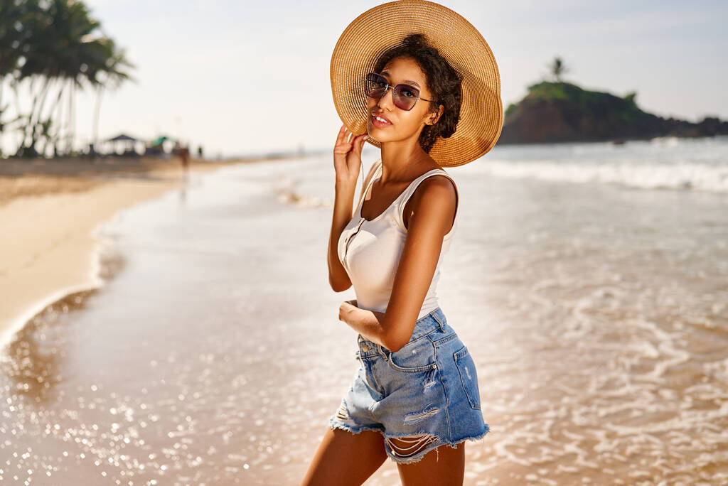 Young african female model in a straw hat and sunglasses posing at resort by sea at sunrise. Black woman against scenic rocky green island and ocean tide at dawn. Multiracial model poses on vacation - Photo, Image