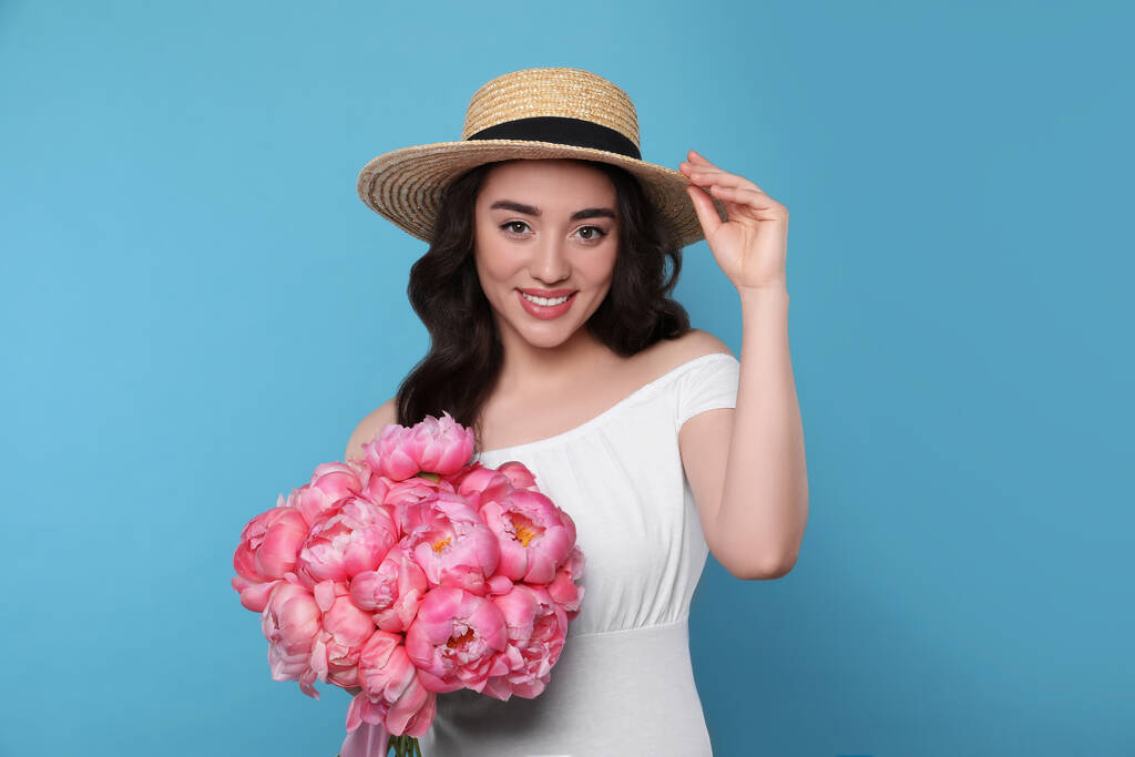 mooie jonge vrouw in stro hoed met boeket van roze pioenen tegen licht blauwe achtergrond - Foto, afbeelding