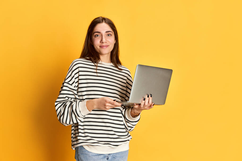 黄色のスタジオの背景にラップトップでポーズ縞模様のセーターの若い笑顔の女の子.就職フェアと学生生活。若者、感情、表情、ライフスタイル、教育、ビジネスの概念 - 写真・画像