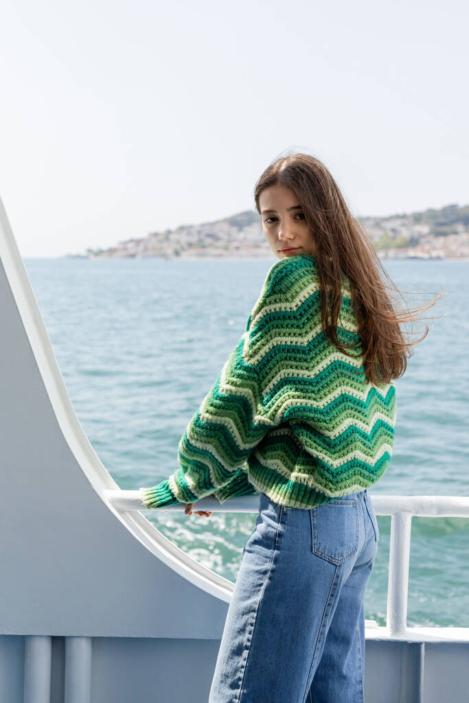 Örgü kazak ve kot pantolon giyen genç bir kadın, arka planda Prenses adalarıyla feribot teknesinde dikilirken kameraya bakıyor. - Fotoğraf, Görsel