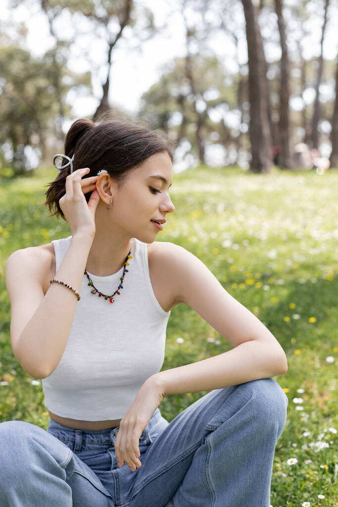 Vue latérale de la jeune femme mettant fleur de marguerite dans les cheveux tout en étant assis sur la pelouse dans le parc  - Photo, image