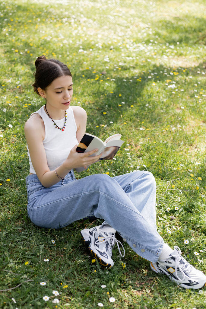 Wysoki kąt widzenia młoda brunetka kobieta w górę i dżinsy czytania książki podczas siedzenia na łące z kwiatami w parku  - Zdjęcie, obraz