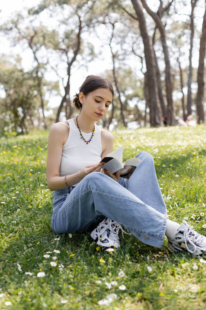 Νεαρή γυναίκα διαβάζει βιβλίο, ενώ κάθεται στο λιβάδι με λουλούδια στο πάρκο  - Φωτογραφία, εικόνα