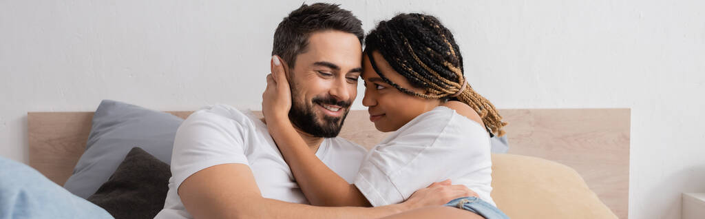 νεαρή Αφροαμερικάνα γυναίκα με ράστα αγκαλιάζει χαρούμενο γενειοφόρο αγόρι στο υπνοδωμάτιο στο σπίτι, πανό - Φωτογραφία, εικόνα