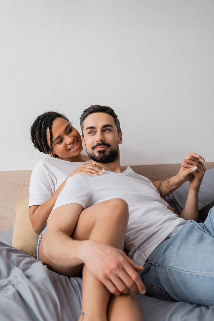 ευτυχισμένο διαφυλετικό ζευγάρι σε λευκό t-shirts κρατώντας τα χέρια, ενώ βρίσκεται στο κρεβάτι στο σπίτι - Φωτογραφία, εικόνα