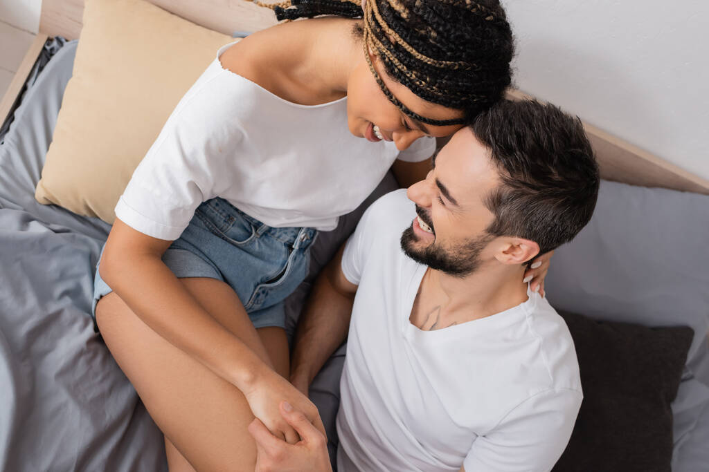vista superior de la pareja interracial despreocupada en camisetas blancas cogidas de la mano y sonriendo en la cama en casa - Foto, Imagen