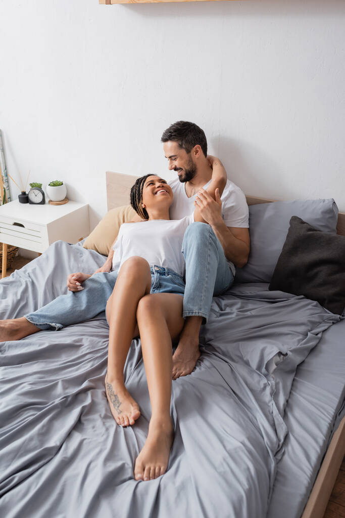 alegre casal multiétnico relaxando na cama em casa e sorrindo um para o outro - Foto, Imagem