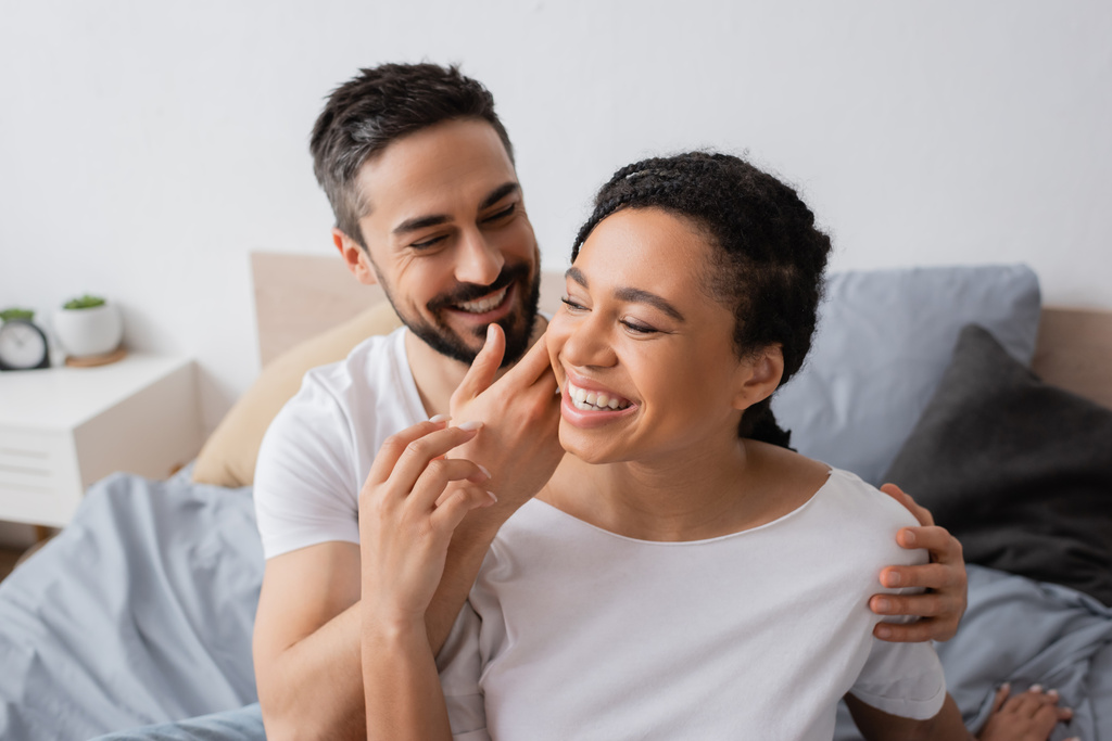 smiling bearded man touching face of overjoyed african american woman in bedroom at home - Photo, Image