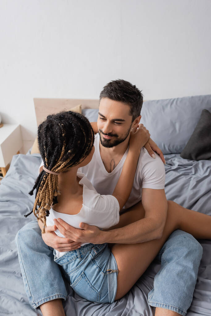 hoge hoek uitzicht van gelukkig bebaarde man en Afrikaanse Amerikaanse vrouw met dreadlocks omarmen op bed thuis - Foto, afbeelding