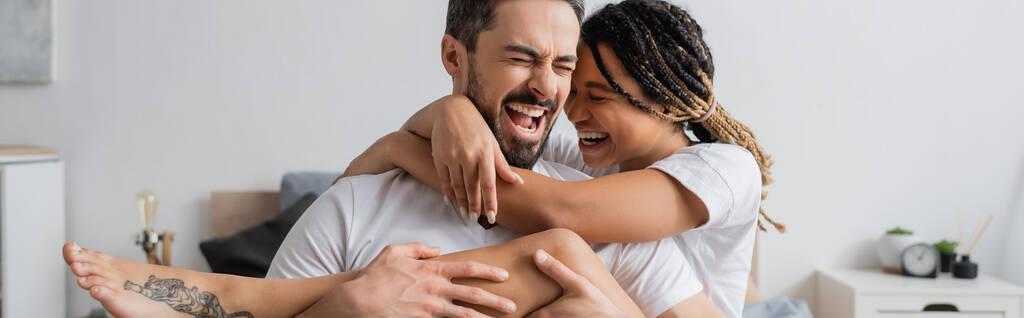 alegre casal interracial em t-shirts brancas abraçando e rindo com os olhos fechados no quarto em casa, banner - Foto, Imagem