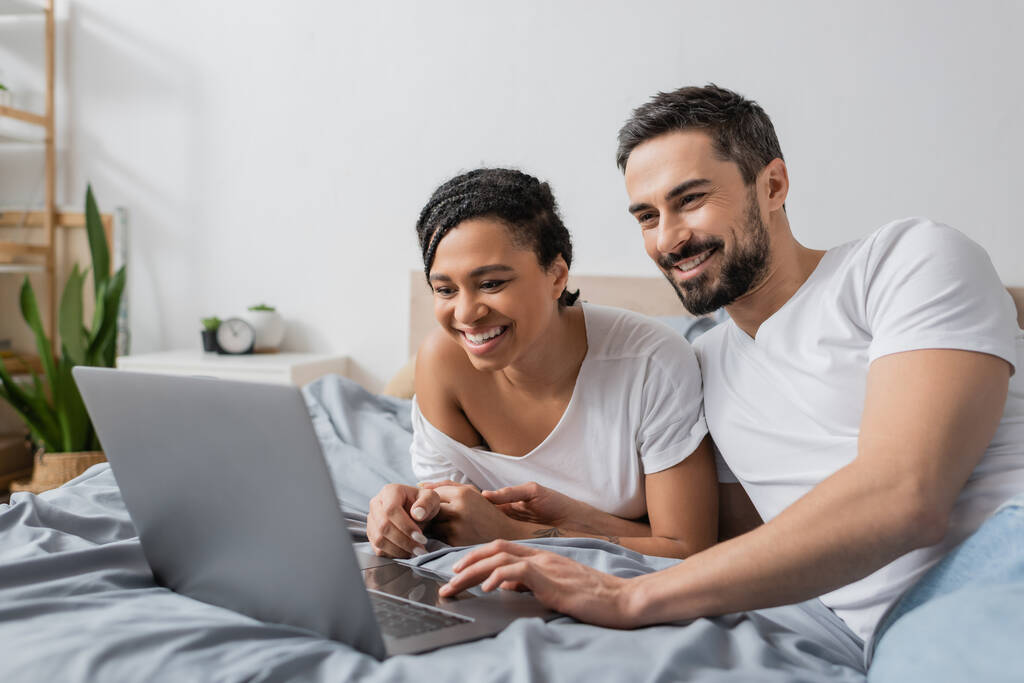 joyful interracial couple in white t-shirts watching movie on laptop in bedroom at home - Photo, Image
