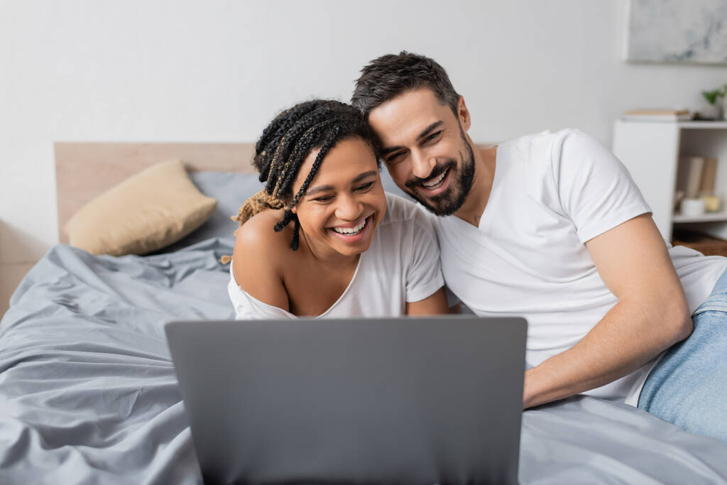 aufgeregtes interrassisches Paar lacht neben Laptop, während es sich zu Hause einen Film auf dem Bett ansieht - Foto, Bild