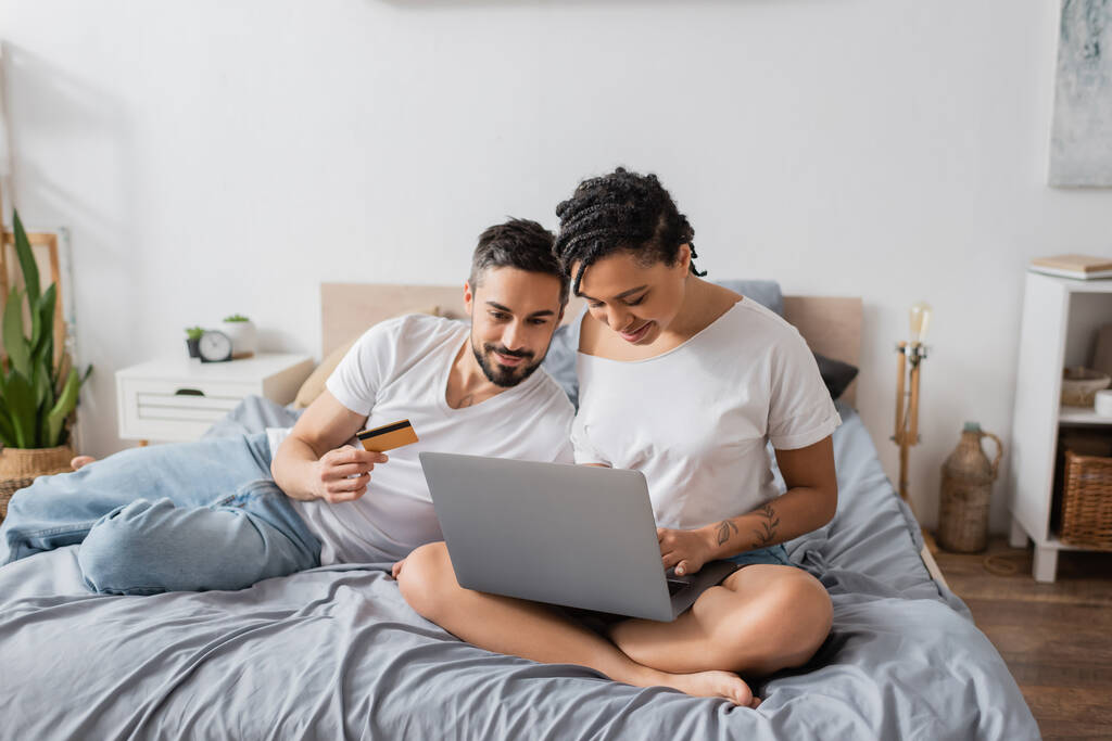 homem barbudo segurando cartão de crédito perto de mulher americana africana sentado com pernas cruzadas e usando laptop na cama em casa - Foto, Imagem