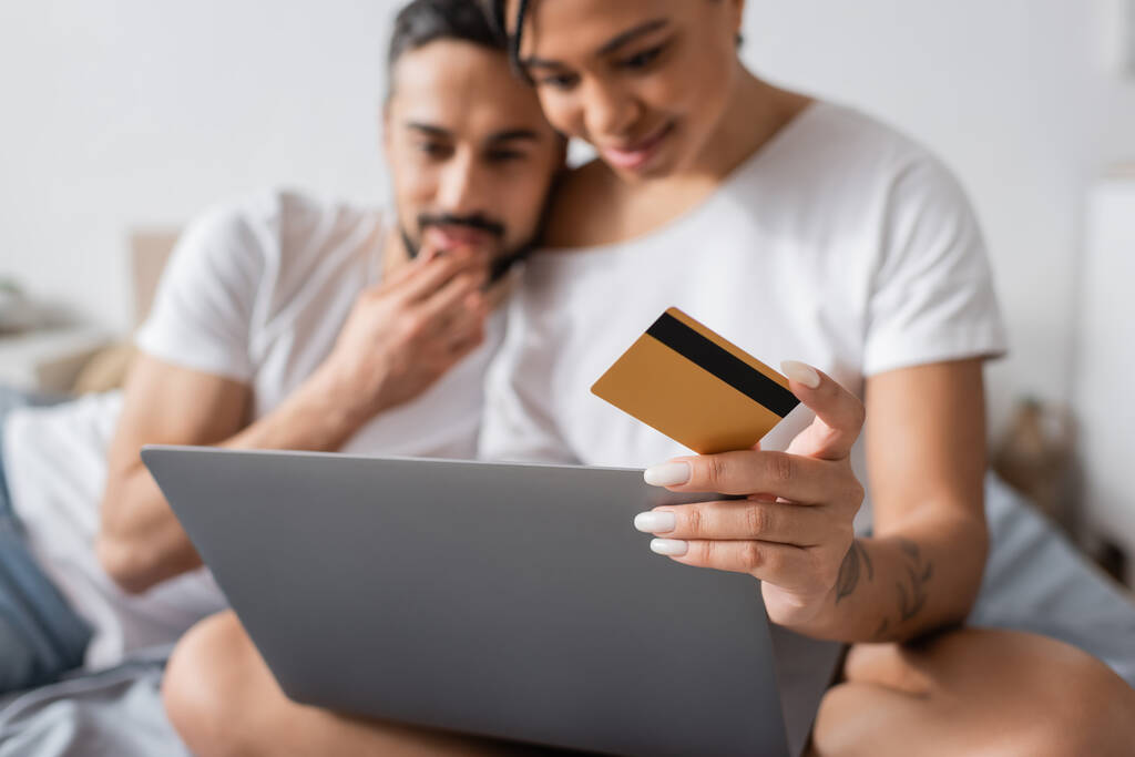 selektywne skupienie się na karcie kredytowej i laptopie w pobliżu rozmytego myślenia międzyrasowego pary podczas zakupów online w sypialni w domu - Zdjęcie, obraz