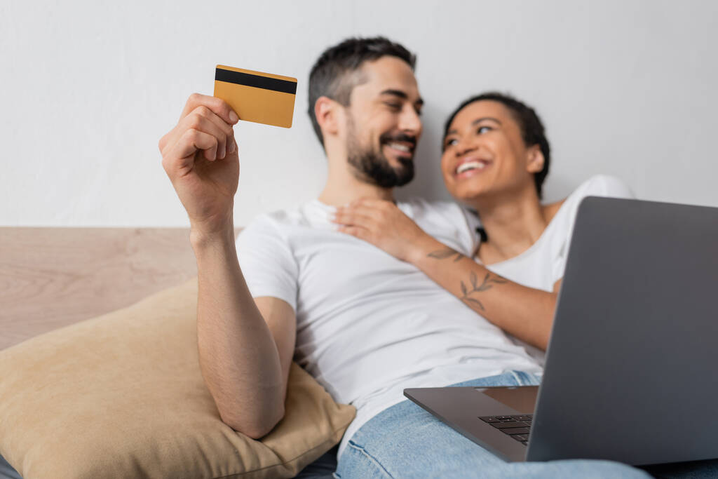 明るい髭の男がノートパソコンの近くにクレジットカードを持っていて幸せなアフリカ系アメリカ人女性が家のベッドルームで - 写真・画像