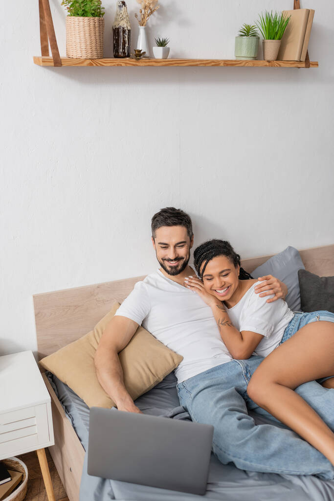 vista de ángulo alto de alegre pareja interracial en camisetas blancas viendo la película en el portátil en la cama en casa - Foto, Imagen