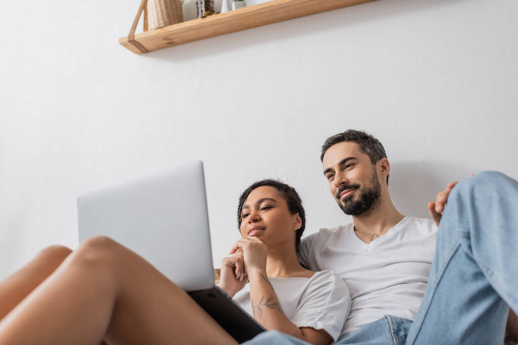 низкий угол зрения улыбающейся многонациональной пары смотреть фильм на ноутбуке во время отдыха в спальне у себя дома - Фото, изображение