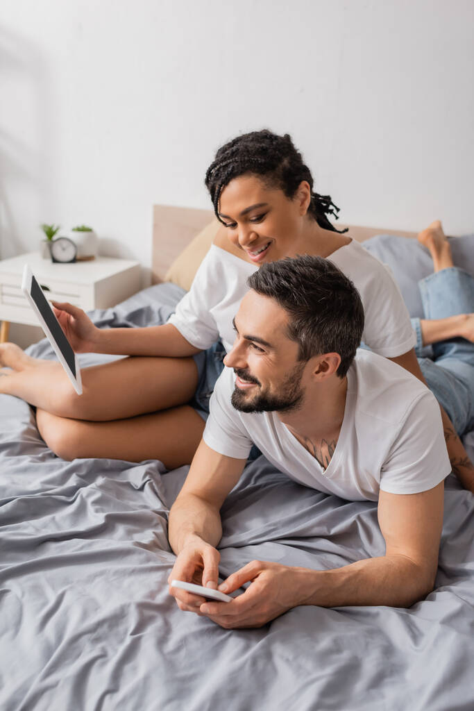 joyful african american woman holding digital tablet near smiling bearded man lying with smartphone on bed at home - Photo, Image