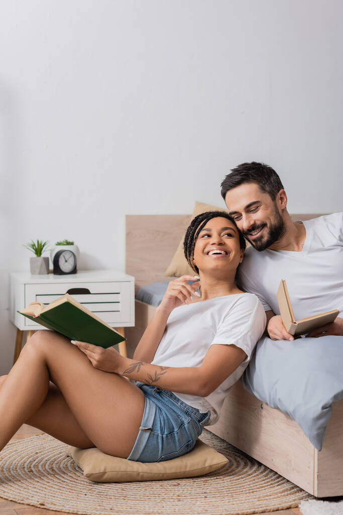 joyful african american woman with book sitting on floor and pillow near bearded boyfriend lying on bed at home - Photo, Image