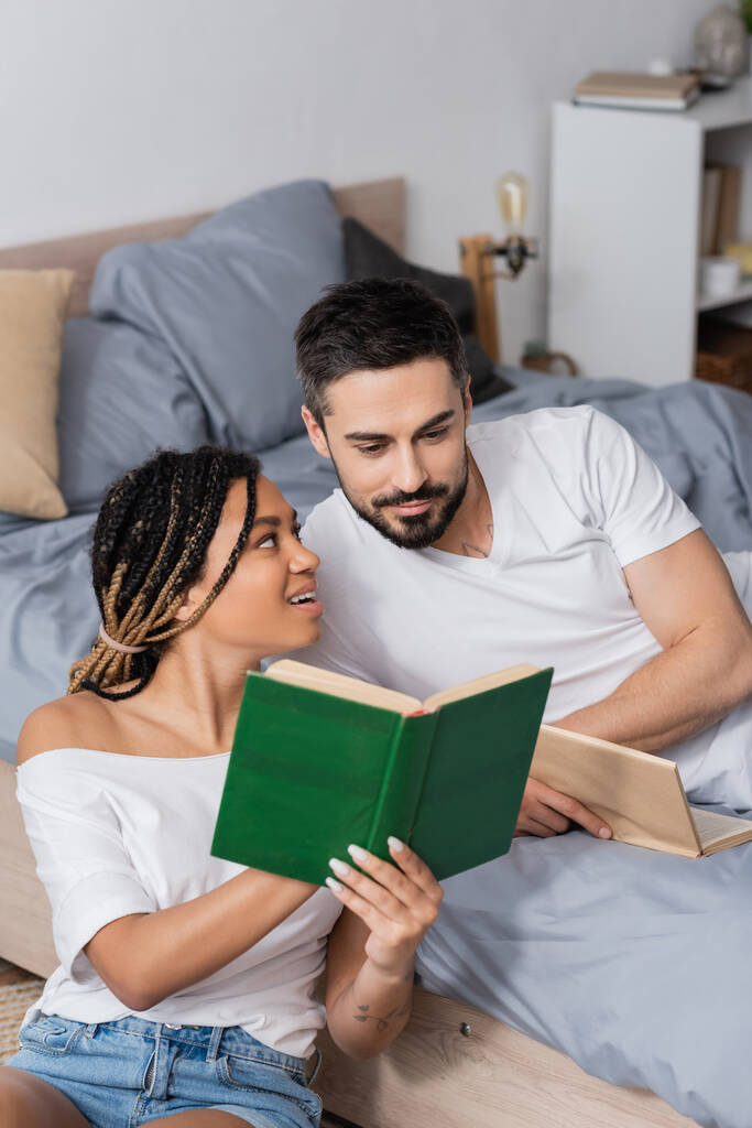 mujer afroamericana sorprendida y feliz mostrando libro a novio sonriente acostado en el dormitorio en casa - Foto, imagen