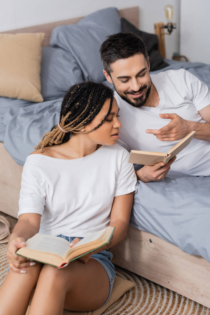 homme barbu souriant pointant vers livre près de jeune femme afro-américaine dans la chambre à coucher à la maison - Photo, image